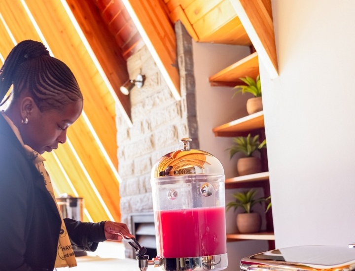 Guest in a cozy wooden lodge pouring bright pink juice from a beverage dispenser on a buffet table with plant-decor shelves nearby.