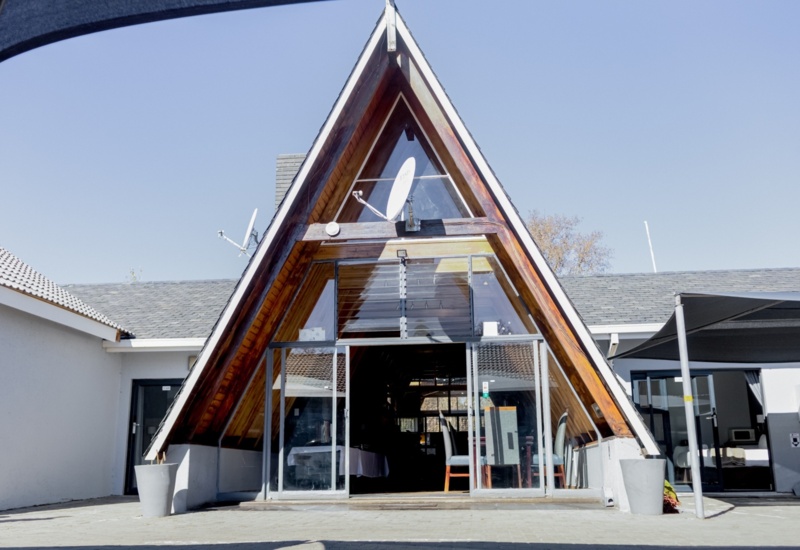 A-frame building with glass entrance, wooden beams, and outdoor seating area at a modern hotel courtyard on a clear day