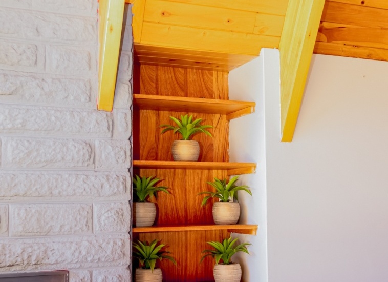 Cozy hotel room corner with light wood ceiling, stone wall, built-in shelves with potted green plants, and white bedding visible
