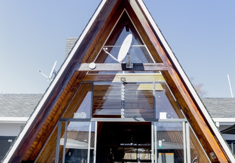 Modern A-frame hotel entrance with wooden beams, glass doors, and seating visible inside under clear blue sky