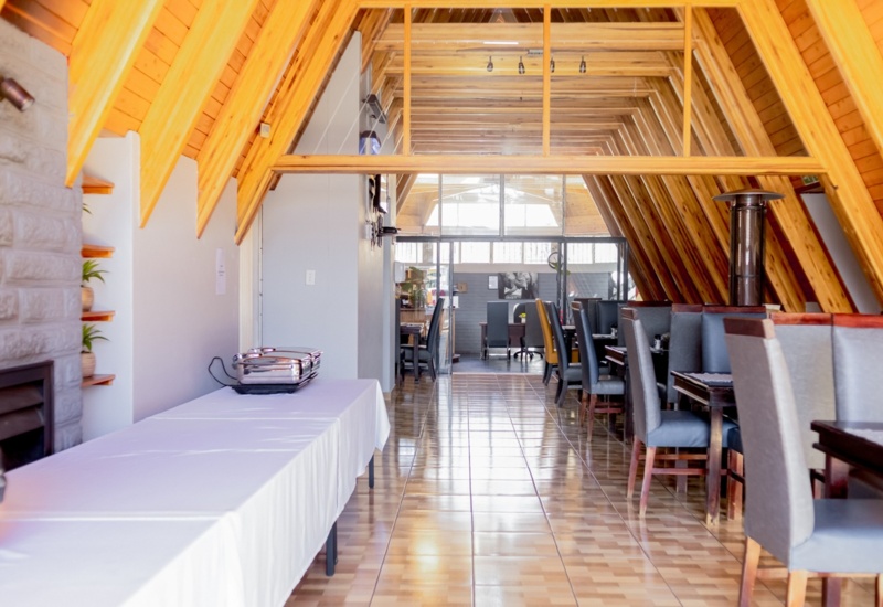 Bright dining area with slanted wooden ceiling, tiled floor, lined tables, plush chairs, and buffet setup with chafing dishes