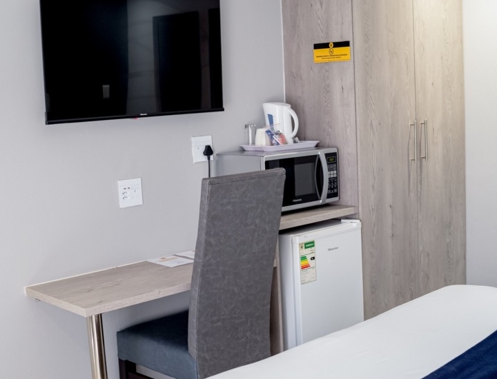 Hotel room with wall-mounted TV, desk and chair, wardrobe, microwave, mini fridge, electric kettle, and a bed with white linens and navy runner