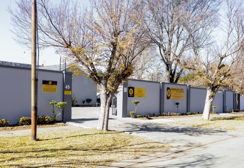 Exterior view of Khayalami Hotel Standerton with gated entrance, gray walls, and leafless trees lining the sidewalk