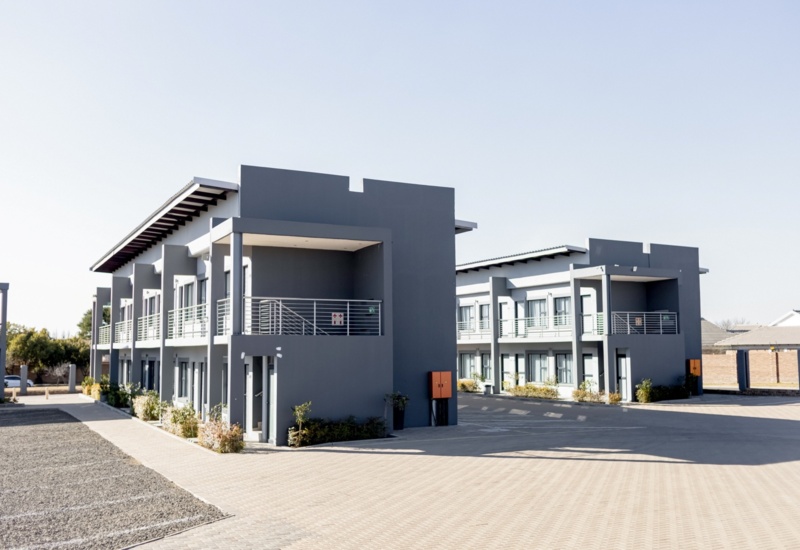 Modern two-story gray hotel buildings with balconies, surrounded by paved walkways and minimal landscaping under clear sky