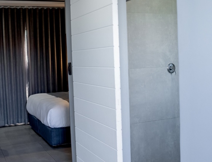 Modern hotel room with a white sliding barn door leading to a gray-tiled shower, bed with white linens, and dark curtains.