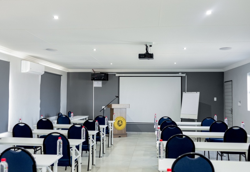 Modern hotel conference room with rows of white tables, navy chairs, bottled water, podium, projector, and pull-down screen