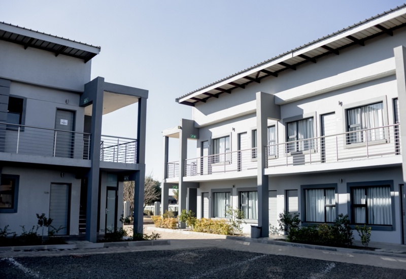 Two-story hotel building with modern design, featuring balconies, large windows, and landscaped shrubbery under clear sky