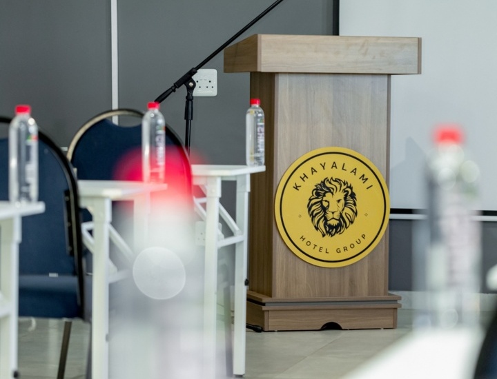 Conference room setup with wooden podium featuring Khayalami Hotel Group logo and microphone, surrounded by chairs and water bottles.