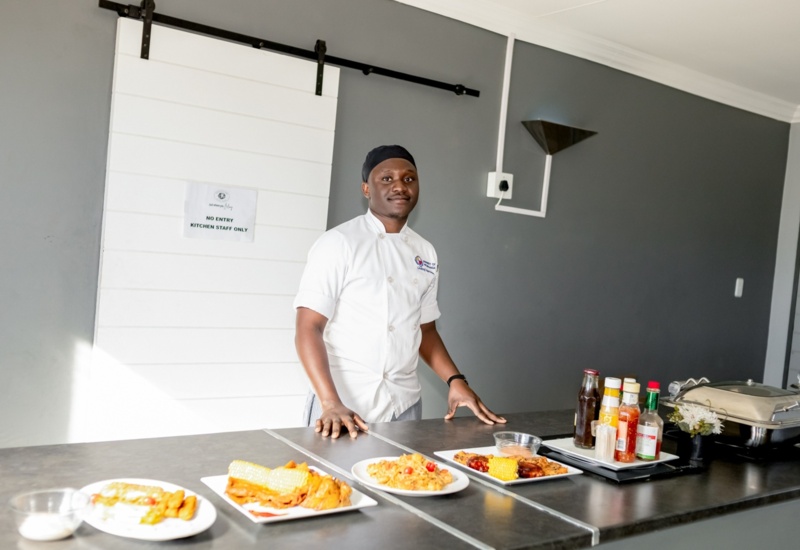 Chef in white uniform standing behind a counter with plated dishes and condiments in a modern hotel kitchen area