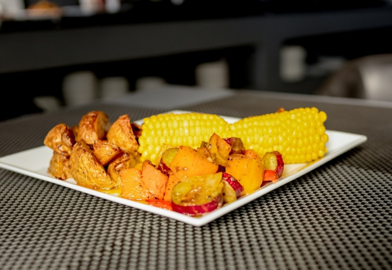 Square white plate with roasted potato wedges, seasoned mixed vegetables, and two bright yellow corn on the cob pieces on a textured table