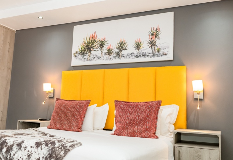 Modern hotel room with bright yellow padded headboard, twin red patterned pillows, soft lighting, and botanical artwork above the bed