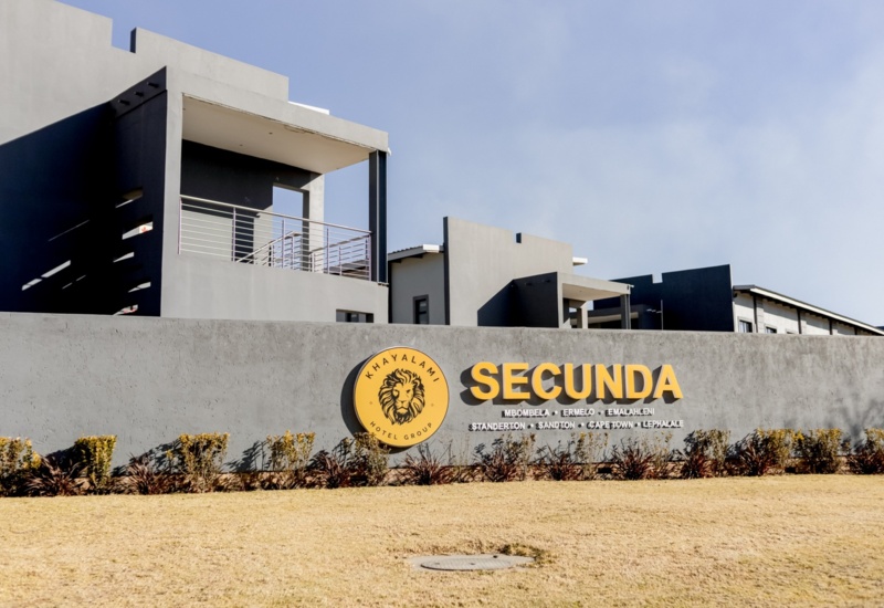 Modern hotel building with gray exterior and balconies behind a wall featuring Khayalami Hotel Group Secunda logo and signage