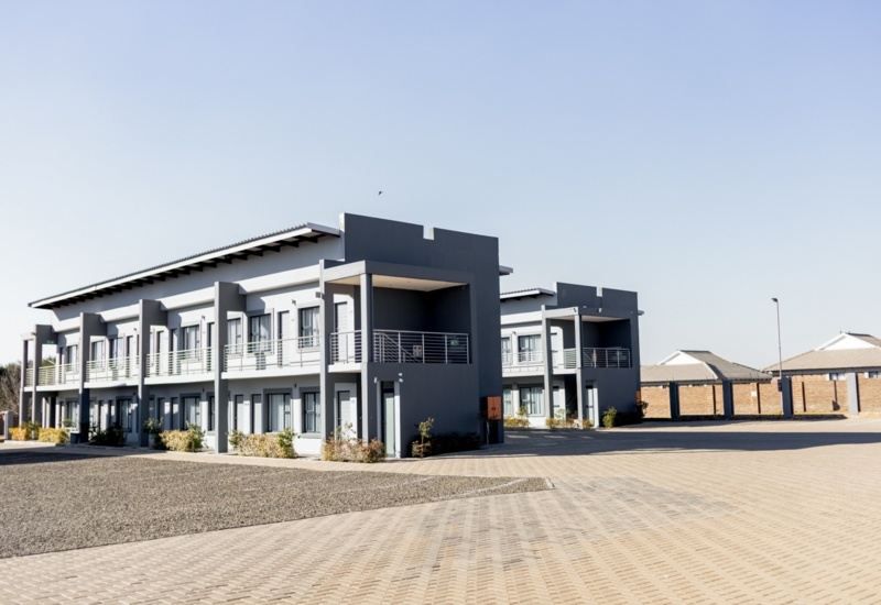 Modern two-story hotel building with multiple guest rooms, balconies, and spacious paved parking area under clear sky