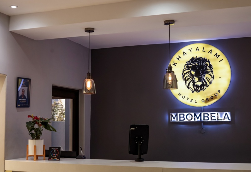 Modern hotel reception with illuminated Khayalami Hotel Group Mbombela sign, pendant lights, and a small plant on white front desk