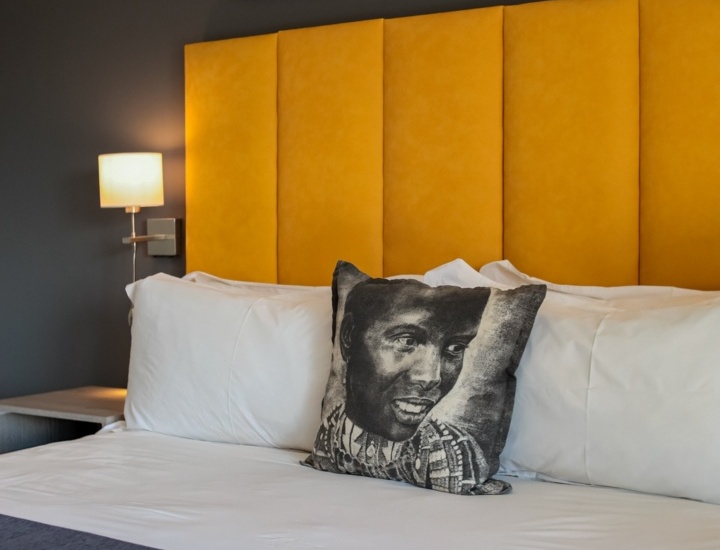 Modern hotel room with a yellow padded headboard, white linens, a decorative pillow with an artistic face, and safari-themed wall art above the bed