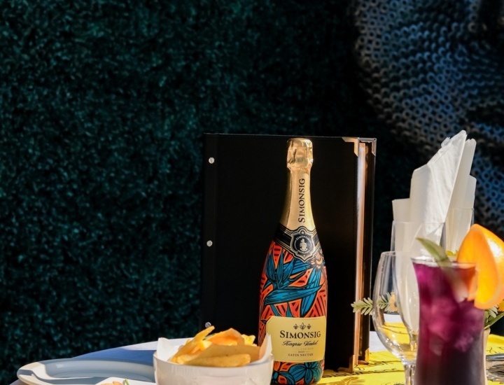 Table set with a colorful bottle of Simonsig wine, a bowl of fries, fried calamari, dipping sauce, and a purple cocktail garnished with an orange slice