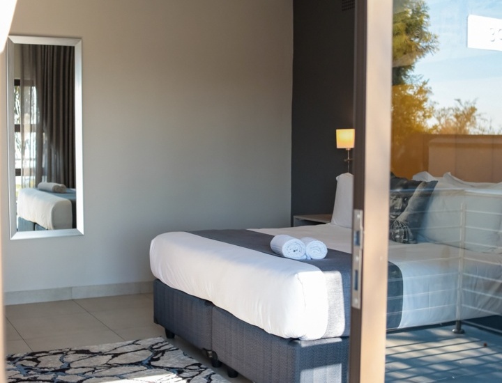 Modern hotel room with a large bed, crisp white linens, rolled towels, a bedside lamp, and a decorative rug on tiled floor