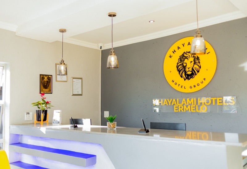 Bright hotel reception with modern gray desk, illuminated blue shelves, pendant lights, and Khayalami Hotels Ermelo logo on the wall