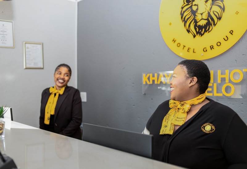 Two smiling hotel receptionists wearing black uniforms and yellow scarves at a modern front desk with a lion logo on the wall