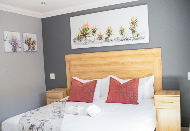 Cozy hotel room with a wooden headboard, white linens, two red accent pillows, matching nightstands, and botanical wall art