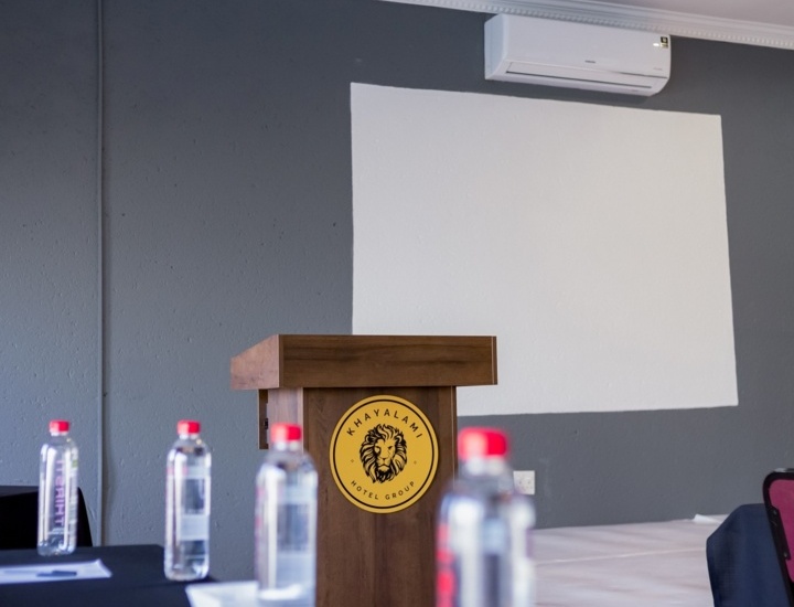 Conference room with wooden podium, branded with lion logo, water bottles on tables, and white projection screen on gray wall