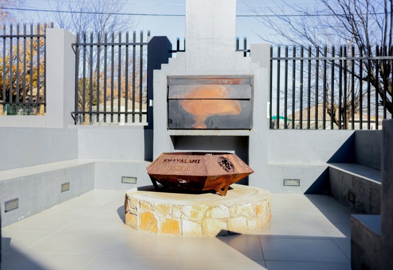 Outdoor fire pit with rusted metal bowl engraved Khayalami Ermelo, surrounded by stone base and concrete bench seating under clear sky