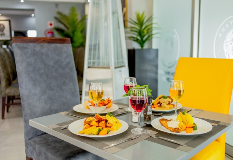 Modern dining table set with plated gourmet meals, assorted wines, and colorful upholstered chairs in bright restaurant ambiance