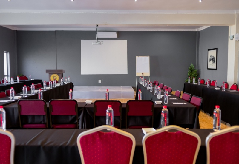 Conference room with U-shaped black tables, red chairs, water bottles, projector screen, podium, and flip chart for meetings or events