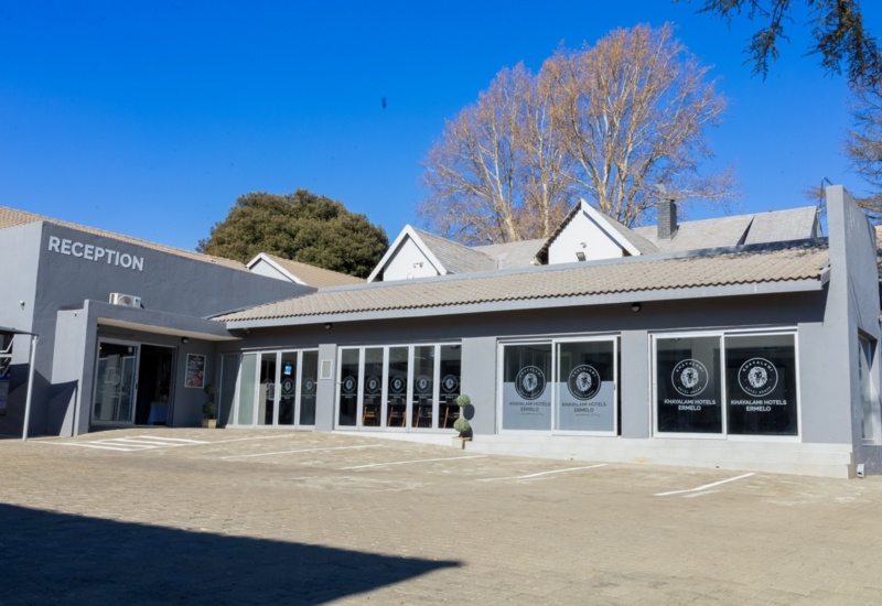 Modern hotel reception building with large glass windows, clear signage, and paved parking area under a bright blue sky