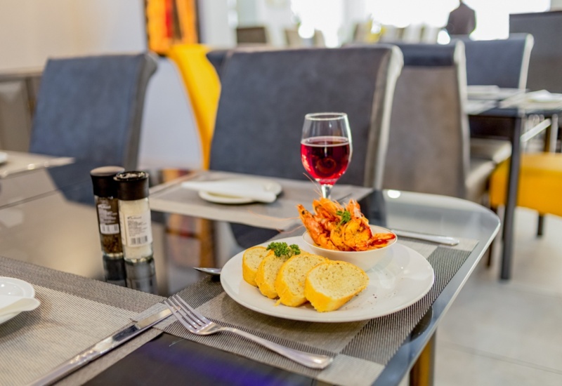 Glass of red wine paired with grilled shrimp and sliced baguette on a modern restaurant table with gray and yellow chairs
