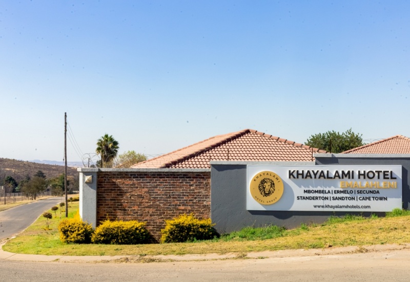 Khayalami Hotel Emalahleni entrance with brick and gray wall, terracotta roof tiles, and landscaped yellow bushes under clear blue sky