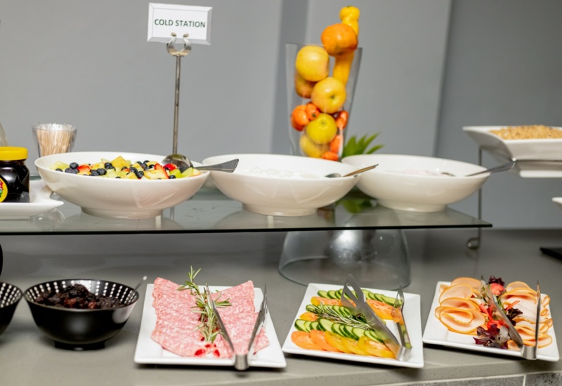 Cold station buffet with fruit salad, sliced meats, fresh cucumber and tomato, yogurt, and whole fruits in a hotel dining setting