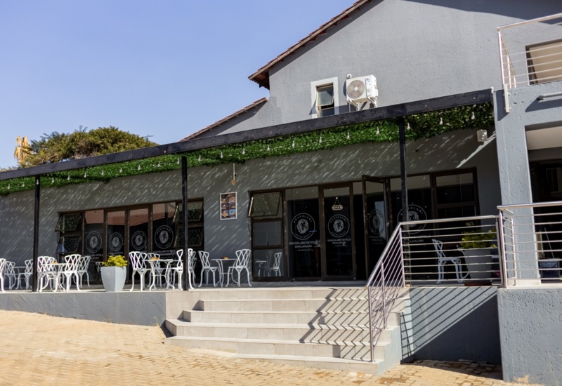 Hotel outdoor terrace with white modern chairs and tables under a shaded pergola with hanging lights, grey building facade