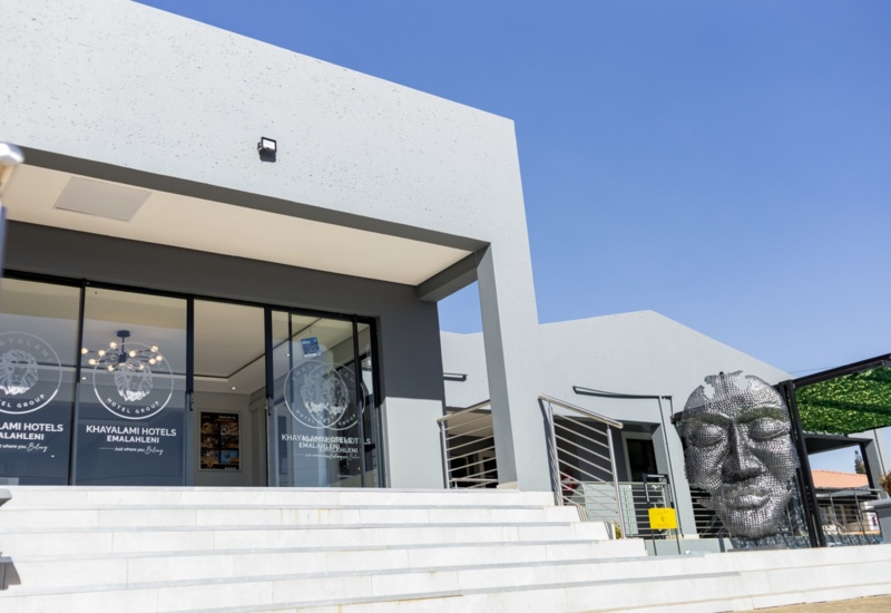 Modern hotel entrance with glass doors, wide white steps, and a large metal face sculpture under a clear blue sky