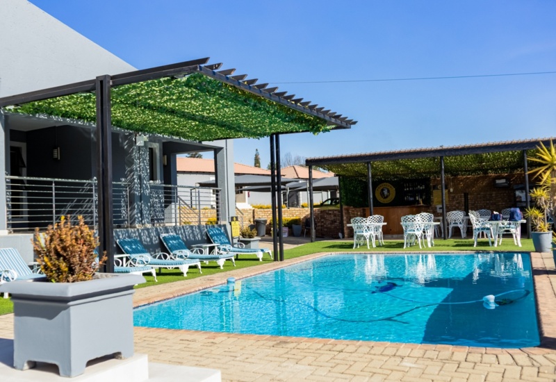 Outdoor hotel pool area with striped lounge chairs under leafy pergolas, white patio tables and bar seating on a sunny day