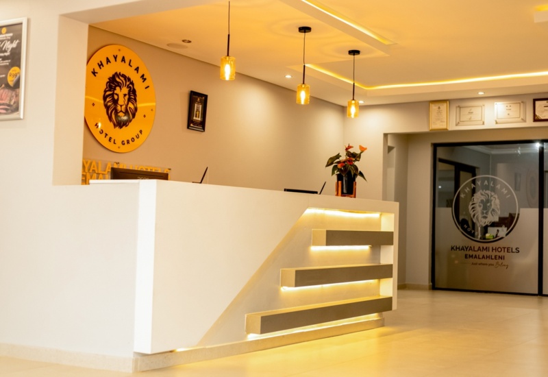 Modern hotel reception with illuminated white desk, warm pendant lights, branded logo on wall, and certificates displayed in the lobby