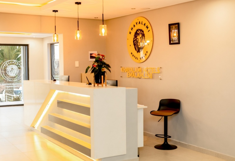 Modern hotel reception with a sleek white desk, warm pendant lights, and the Khayalami Hotels logo on a beige wall