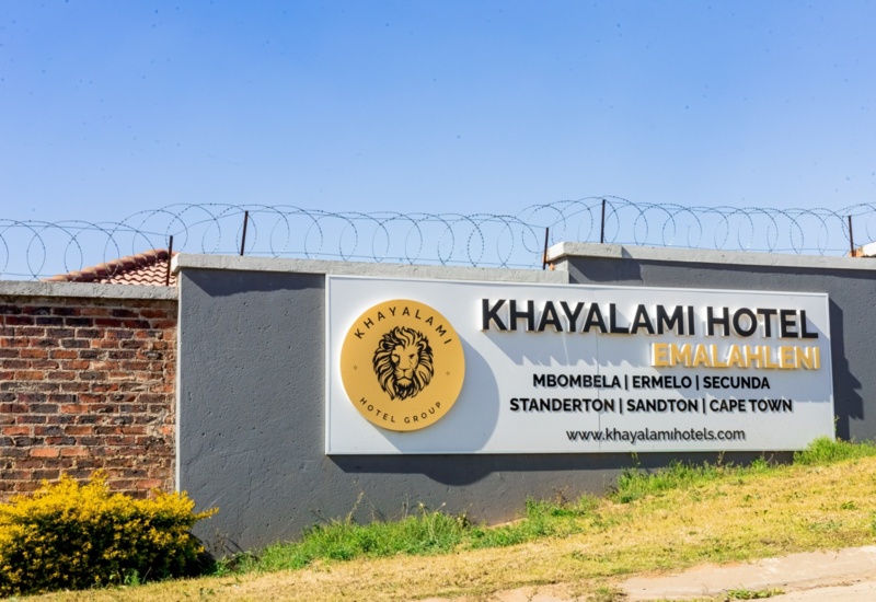 Exterior hotel sign on grey wall with barbed wire showing Khayalami Hotel Emalahleni and locations including Cape Town and Sandton
