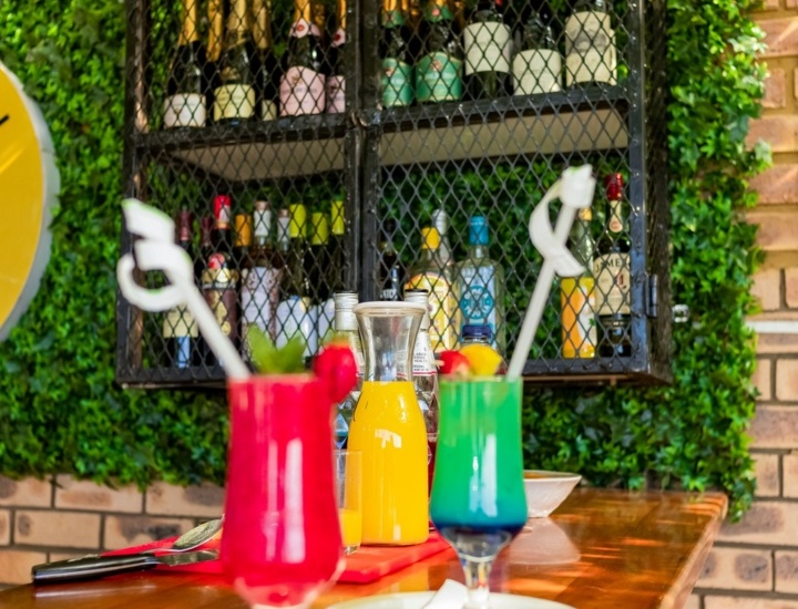 Colorful cocktails and orange juice served on a wooden bar counter with a green ivy wall and wine bottles in a black metal rack