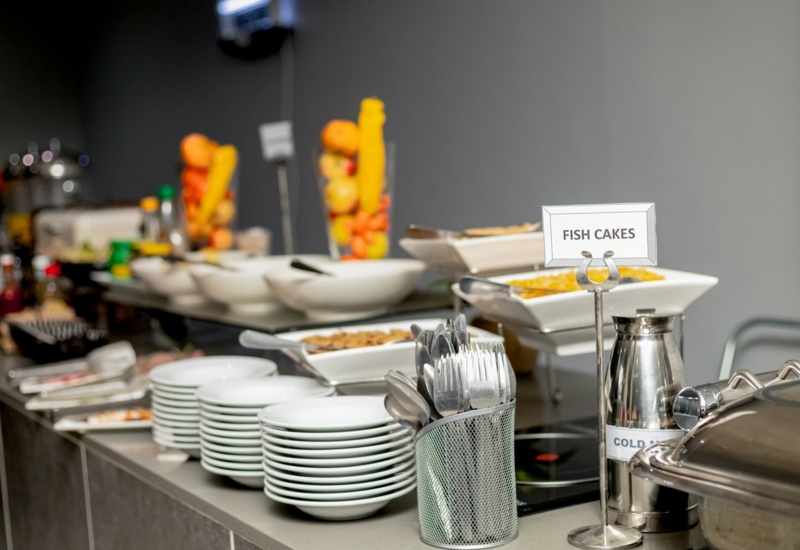 Buffet table setup with stacks of white plates, cutlery holder, dishes labeled fish cakes, and various condiments in a modern dining area
