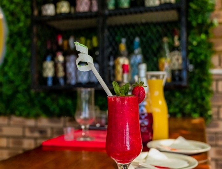 Vibrant red berry cocktail garnished with mint and strawberry, placed on a white plate at a wooden bar with bottles in the background