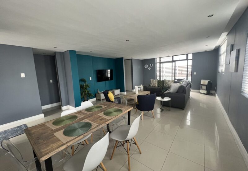 Spacious living and dining area with a wooden table, modern chairs, blue accent wall, flat-screen TV, and cozy seating near large windows