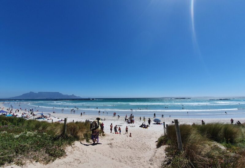Sunny beach with white sand and people swimming, sunbathing, and playing, with a distant view of a flat-topped mountain under clear blue sky