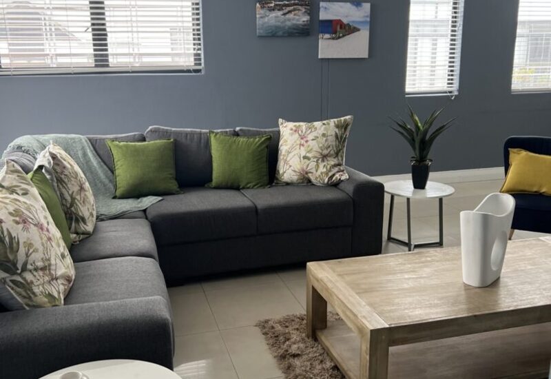 Modern hotel lounge with gray sectional sofa, floral and green pillows, wood coffee table on shag rug, and natural light from large windows