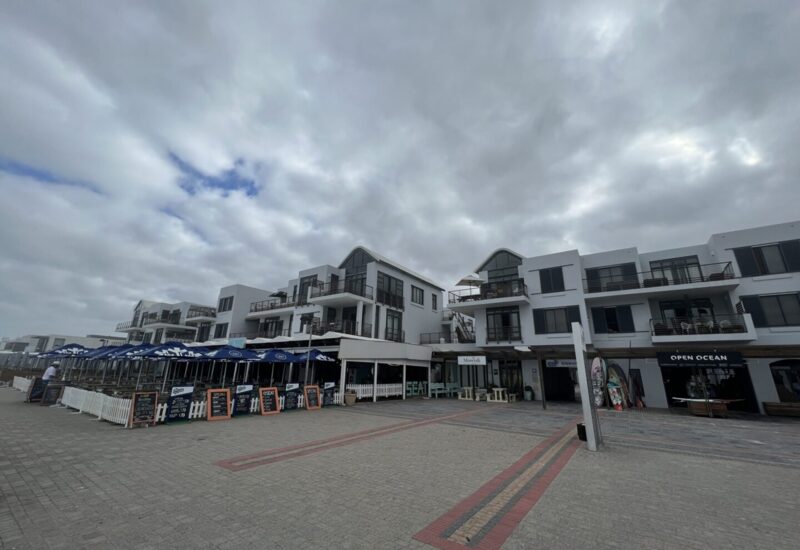 Coastal modern white hotel complex with multiple balconies, outdoor dining under blue umbrellas, and beachfront shops on a cloudy day