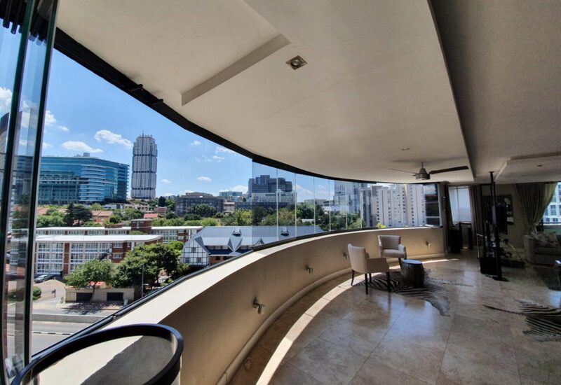 Spacious hotel balcony with curved glass railing offering panoramic city skyline views and two armchairs for relaxation