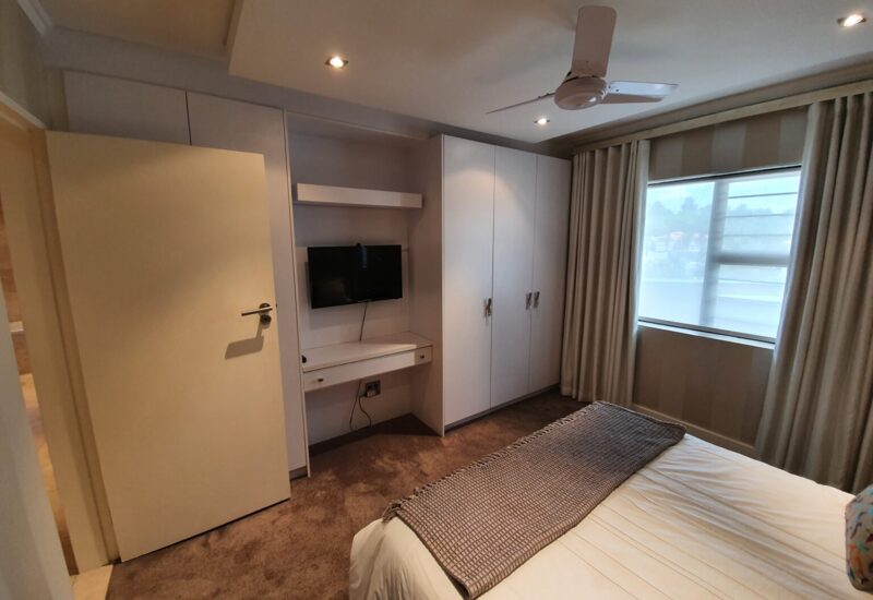 Cozy hotel bedroom with a double bed, textured throw, built-in white wardrobes, wall-mounted TV, ceiling fan, and a window with beige curtains