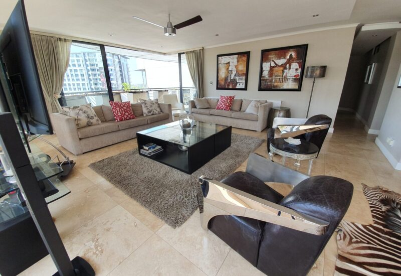 Modern hotel suite living area with beige sofas, red accent pillows, black leather chairs, glass coffee table, and balcony with city view