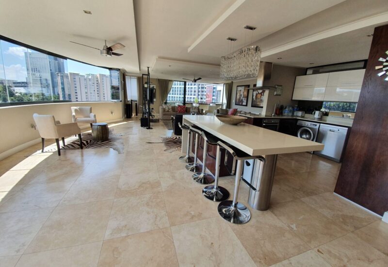 Spacious open-plan suite with modern kitchen island, bar stools, living area, large windows offering cityscape views, and natural light