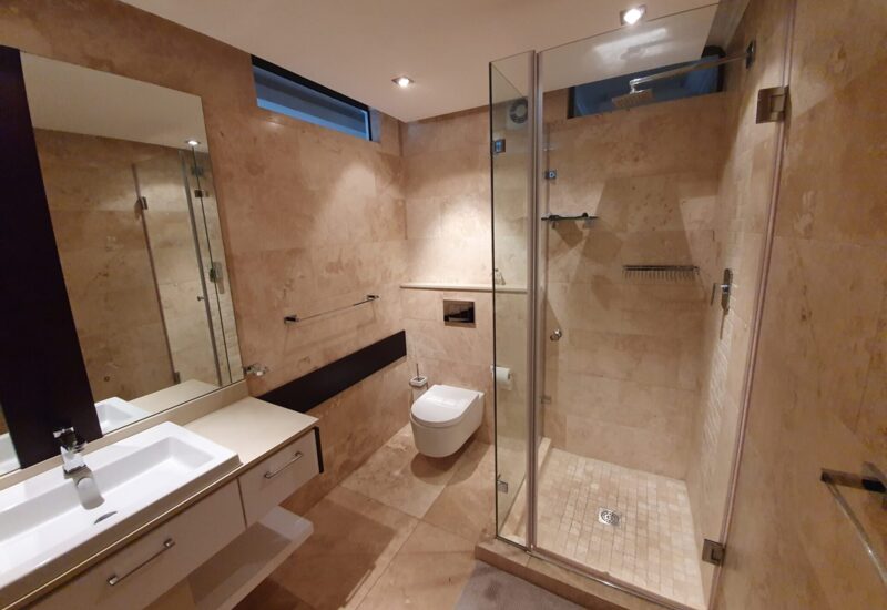 Modern hotel bathroom with beige stone tiles, glass-enclosed shower, wall-mounted toilet, and sleek white sink vanity
